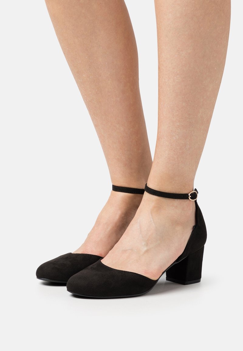 Jambes portant des escarpins noirs en daim à bout fermé avec talons blocs, fines brides autour de la cheville et boucle dorée sur fond blanc.