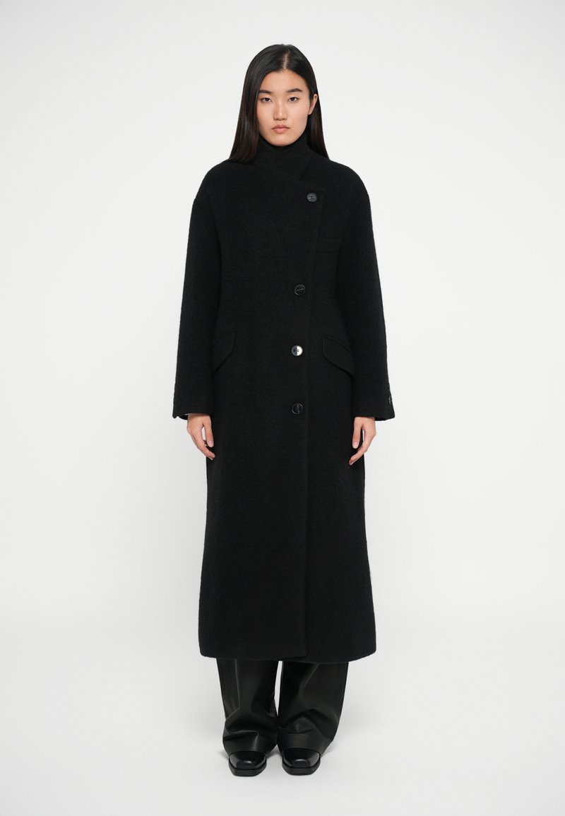 Melna vilnas mēteli ar augstu apkakli, kas aprīkots ar vienu pogu, sānu kabatām un garām piedurknēm, apvienots ar melnajiem biksēm un apaviem.