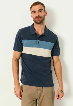 Mann trägt ein kurzärmeliges marineblaues Polohemd mit blauen und weißen Streifen, steht vor einem einfarbigen hellen Hintergrund.