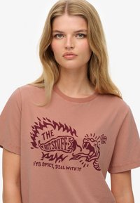 Camiseta de manga corta en rosa empolvado con un cuello redondo que presenta un estampado gráfico en rojo oscuro de llamas y una serpiente. El texto dice "¡LO CALIENTE! ES PICANTE, ¡SUPÉRALO!"