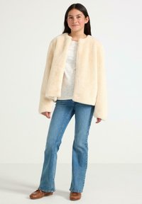 Creme-kleurige nepbontjas met een ronde kraag, oversized pasvorm en verlaagde schouders, gecombineerd met blauwe flared jeans en bruine schoenen.