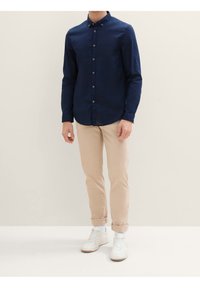 Camisa de botones de manga larga en azul marino con collar y acentos en los botones en contraste, combinada con pantalones beige remangados y zapatillas blancas.