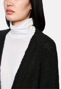 Schwarze, strukturierte Strickjacke über einem weißen Rollkragenpullover, die einen weichen, flauschigen Stoff mit offenem Frontdesign und subtilen Details präsentiert.