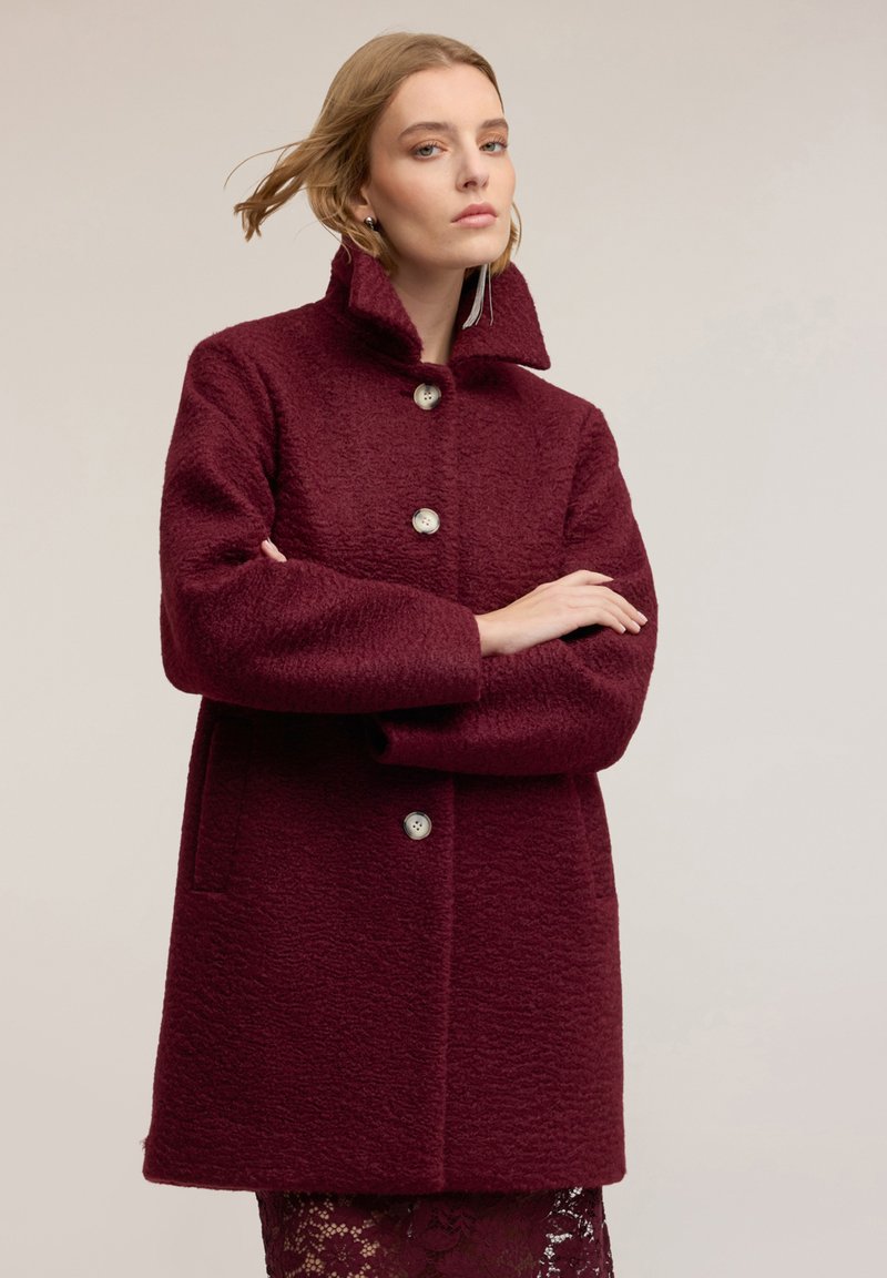 Bordó kabát s texturou a vysokým límcem, vybavený dvěma knoflíkovými zapínáními, dvěma bočními kapsami a volným střihem.