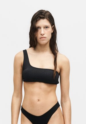 Jeune femme aux cheveux noirs mouillés portant un haut de bikini texturé noir à une épaule et un bas de bikini taille haute assorti, debout devant un fond clair uni.