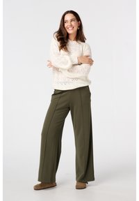 Pull en crème tricoté avec un motif de cœur, pantalons olive à jambes larges et chaussons marron. Le mannequin pose les bras croisés, souriant contre un fond blanc.