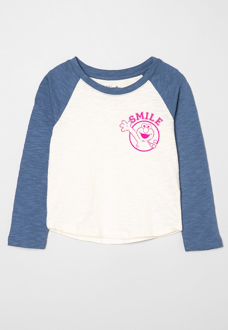 T-shirt à manches raglan longues avec des manches bleues et un corps crème. Présente le mot "SMILE" et un graphique de personnage en broderie rose sur le devant. Texture de tissu douce.