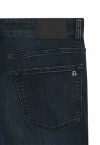 Jeans de denim oscuro con un parche de cuero en la cintura y un bolsillo trasero con un logo en relieve. Costuras simples y diseño limpio.