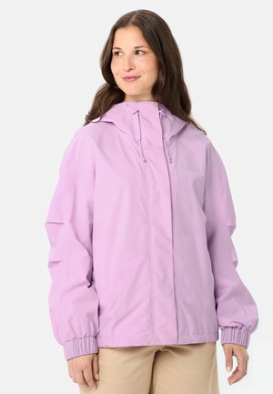 Frau mit langen braunen Haaren, die eine hellviolette Kapuzenjacke und beige Hose trägt, blickt nach links mit einem leichten Lächeln.