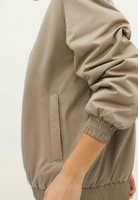 Veste bomber beige avec poignets et ourlet élastiques. Tissu lisse, poche latérale visible. Design simple et arrondi, sans motifs ni ornements.