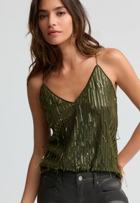Olivgrünes Camisole mit V-Ausschnitt, ausgestattet mit vertikaler Fransenverzierung und Pailletten. Dünne Schulterträger, geschichtete Textur. Schwarze Hosen.