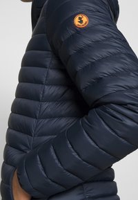 Giacca imbottita blu navy con design trapuntato, che mostra una texture liscia e una patch in gomma sul braccio superiore. Caratterizzata da tasche laterali con zip.