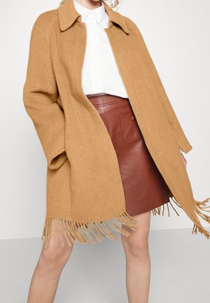 Femme portant un manteau camel à franges par-dessus une chemise blanche à col et une jupe en cuir marron avec une poche avant et un bouton.