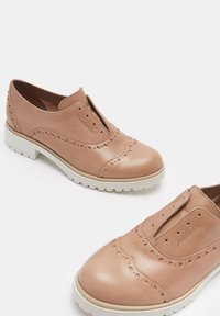 Chaussures brogue en cuir tan à enfiler, avec des perforations décoratives et des semelles en caoutchouc blanches sur fond blanc.