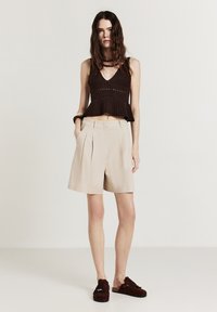 Donna che indossa un top senza maniche in maglia marrone scuro, pantaloni corti plissettati beige a vita alta, scarpe scivolate marrone scuro e gioielli minimal.