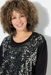 Felpa nera con un grande dettaglio di paillettes nere e argentate sul davanti. Capelli ricci e trucco minimal completano il design.