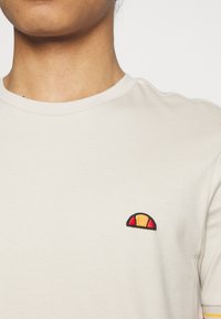 Gros plan sur un torse portant un t-shirt beige avec un petit logo en demi-cercle rouge et orange sur la poitrine gauche.
