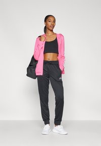 Różowa bluza z zamkiem, czarny top crop, czarne dresy z ściągaczami na kostkach i białe sneakersy. Model trzyma czarną torbę sportową.