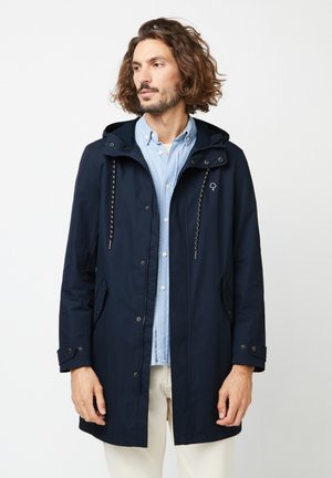 Homme portant un manteau à capuche bleu marine par-dessus une chemise bleu clair à boutons et un pantalon blanc, debout devant un fond blanc uni, regardant vers la gauche.