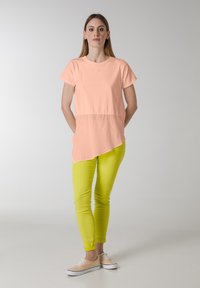 Ferskenfarvet asymmetrisk t-shirt med en blød tekstur, parret med slim-fit klare gule bukser og lyse beige sneakers.