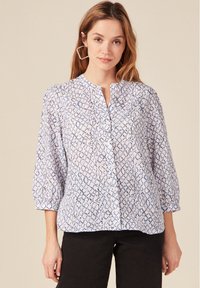 Blusa bianca leggera e semi-trasparente con motivo astratto blu, chiusura a bottoni e maniche a 3/4, abbinata a pantaloni scuri.