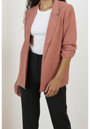 Kobieta ubrana w różowy marynarkę w odcieniu pudrowego różu z podwiniętymi rękawami, biały prążkowany top oraz czarne spodnie, stojąca na jednolitym tle.