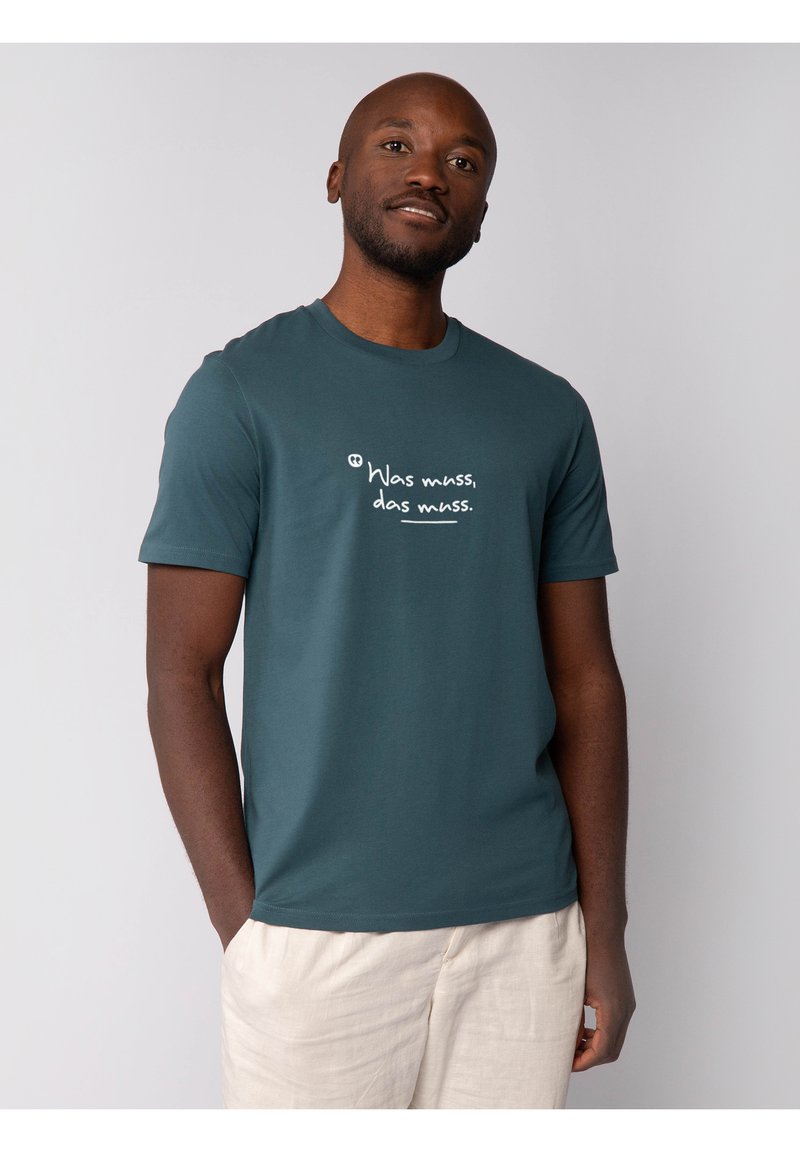 Teal katoenen T-shirt met een ronde hals en korte mouwen. Heeft witte opdruk op de voorkant. Geloofd met lichtgekleurde, losse broek.
