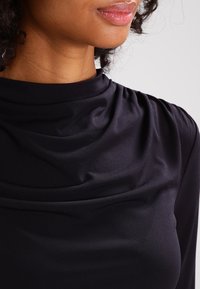 Svart långärmad topp med hög halsringning, med draperat tyg runt kragen och slät textur, lämplig för olika tillfällen.