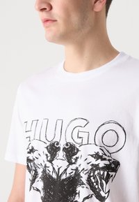 Hvit bomulls t-skjorte med en dristig svart grafikk som viser to ulver og ordet "HUGO" trykket over. Klassisk rund hals, korte ermer.