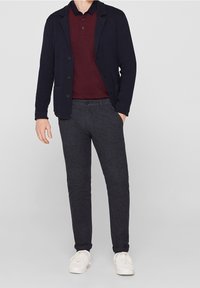 Marineblauwe cardigan over een bordeauxrode polo, gecombineerd met grijze textuurbroek en witte sneakers. De cardigan heeft knopen en heeft een casual pasvorm.