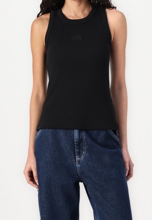 Mujer con top negro sin mangas y vaqueros azules sueltos de cintura alta, de pie con las manos relajadas a los lados.