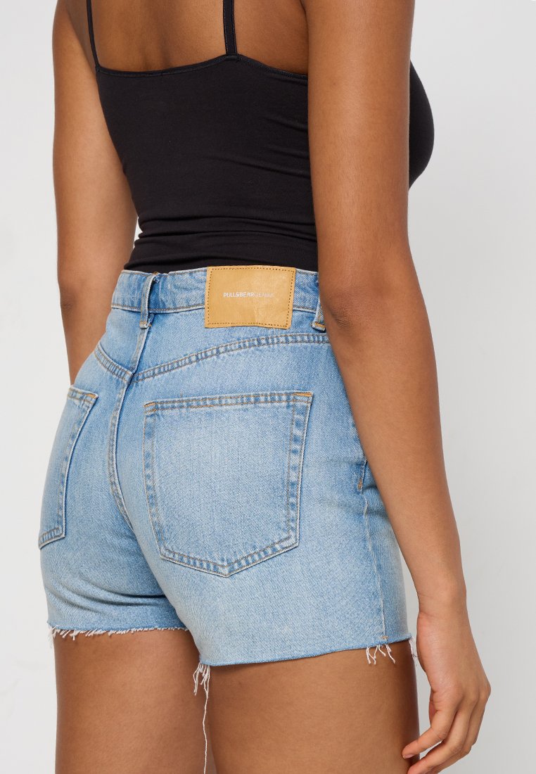 Pantalones cortos de mezclilla azul claro con cintura alta, dobladillo deshilachado y bolsillos traseros. Presenta un parche de cuero marrón claro en la cintura.