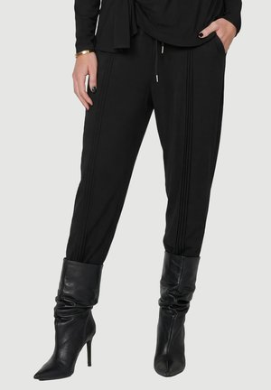 Donna che indossa pantaloni neri aderenti con dettagli di cuciture verticali e stivali neri larghi con punta a punta e tacco alto, mani in tasca.