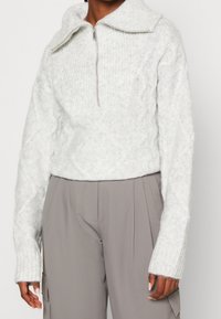 Ingekorte lichtgrijze trui met een hoge kraag, een halve ritssluiting aan de voorkant en een gestructureerd kabelgebreid patroon. Gecombineerd met losse, beige broek.