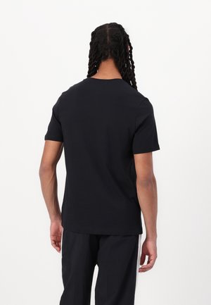 Persona con cabello largo y rizado lleva una camiseta negra lisa de manga corta y pantalones negros, de espaldas sobre un fondo blanco.