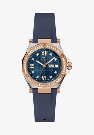 Oktagona pulksteņa ar tumši zilu gumijas siksniņu, rozā zelta akcentiem, zilā teksturētā ciparnīcā, kristāla stundu marķieriem un datuma rādītāju.