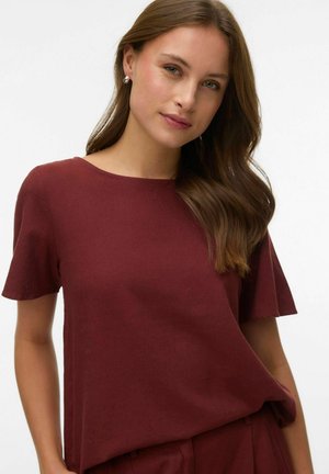 Jeune femme aux longs cheveux bruns portant des boucles d'oreilles argentées et un haut bordeaux à manches courtes, se tenant devant un fond blanc.