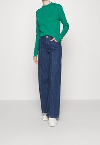 Maglietta verde a maniche lunghe con scollatura rotonda, abbinata a jeans blu in denim a gamba larga e vita alta e sneakers bianche. Design semplice e texture.