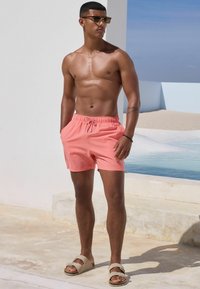 Rosa karierte Badeshorts mit einem Kordelzugbund, kombiniert mit beigen Sandalen. Im Hintergrund sind ein Pool und minimalistische Architektur zu sehen.