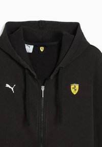 Sort hættesvetrt med lynlås lavet af blødt stof, der har et hvidt Puma-logo og et gult Ferrari-emblem med en farvet accent.