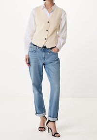 Béžová vesta s černými knoflíky přes bílou košili, spojená s modrými zkrácenými džíny a černými sandály na vysokém podpatku.