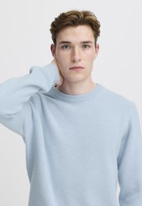 Svetlo modra pulover s čolnasto izrezom, narejen iz mehke tkanine. Ima rebraste manšete in izrez. Model ima kratke rjave lase in nevtralen izraz na obrazu.