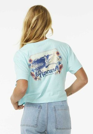 Vrouw met lang blond haar die een lichtblauw Ripcurl T-shirt draagt met een surfer- en bloemmotief, gecombineerd met hoogwaardige spijkerbroeken.