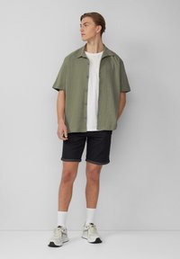 Chemise olive verte à manches courtes avec un col, t-shirt blanc en dessous, shorts sombres retroussés et baskets blanches sportives.