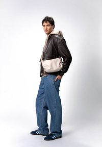 Giacca di pelle marrone, felpa beige con cappuccio, jeans blu larghi e scarpe blu navy con strisce bianche. Gli accessori includono una borsa a tracolla beige chiaro.