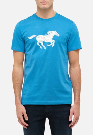 Mężczyzna w jasnoniebieskiej koszulce z krótkim rękawem z białym graficznym motywem galopującego konia na piersi, w połączeniu z ciemnymi dżinsami.