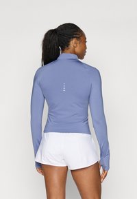 Long-sleeve, light purple athletic top with a high collar, fitted design, and a back accent of five vertical lines. Paired with white shorts.