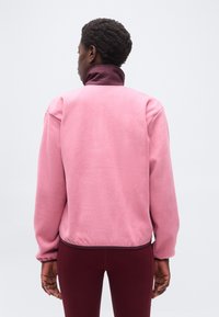 Άτομο με κοντά σγουρά μαλλιά που φοράει ροζ ζεστό μπουφάν με ψηλό γιακά και μπορντό παντελόνι, στέκεται με την πλάτη στραμμένη.