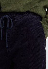 Pantalons en velours côtelé bleu marine avec une surface texturée et une taille élastique, dotés d'un cordon de serrage et associés à un pull vert olive.
