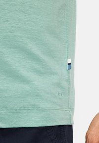 Minzgrünes T-Shirt mit kurzen Ärmeln aus weicher Baumwolle, mit einem seitlichen Saumakzent, der weiße und navyblaue Streifen zeigt. Dezente Textur und Logodetail.